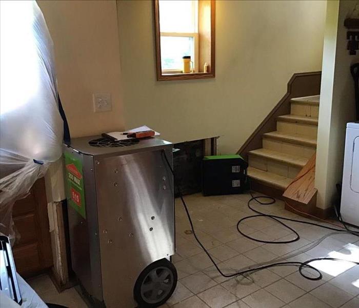 Silver and green dehumidifier on a tile floor with a stairway and a green air mover on the floor.